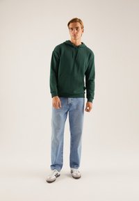 Hanorac verde din bumbac cu buzunar frontal și glugă cu șnur, asortat cu blugi denimu de un bleu deschis și adidași albi cu accente negre.