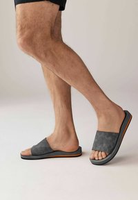 Harmaat mokkasaappaat, joissa on leveä remmi, teksturoitu pinta ja oranssi yksityiskohta anturan alueella. Matalaprofiilinen design.