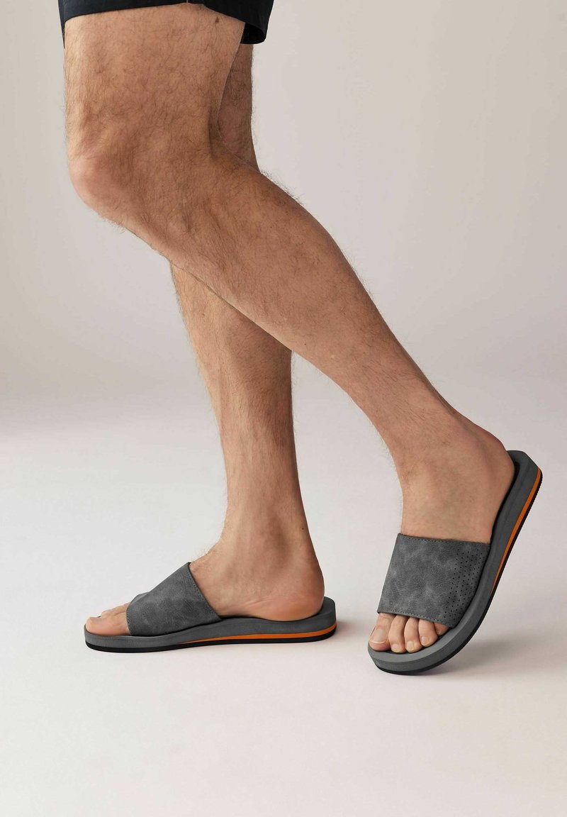 Harmaat mokkasaappaat, joissa on leveä remmi, teksturoitu pinta ja oranssi yksityiskohta anturan alueella. Matalaprofiilinen design.