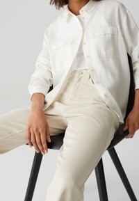 Chemise blanche boutonnée avec deux poches poitrine, portée sur un débardeur blanc. Associée à un jean taille haute beige clair.