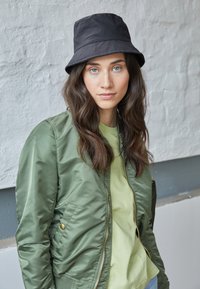 Casaco bomber verde com uma textura brilhante, fecho de correr na frente e hardware dourado. Por baixo, uma t-shirt verde clara. Um chapéu tipo balde preto completa o visual.