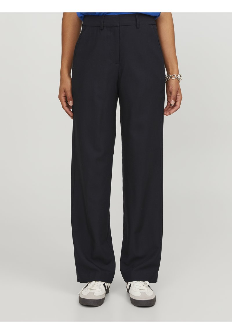 Personne portant un pantalon noir à jambes droites et des baskets blanches, se tenant devant un fond uni clair.