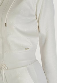 Felpa bianca con zip, realizzata in tessuto testurizzato, dotata di hardware in ottone e motivi in rilievo sottili, con vita elasticizzata e cordino.