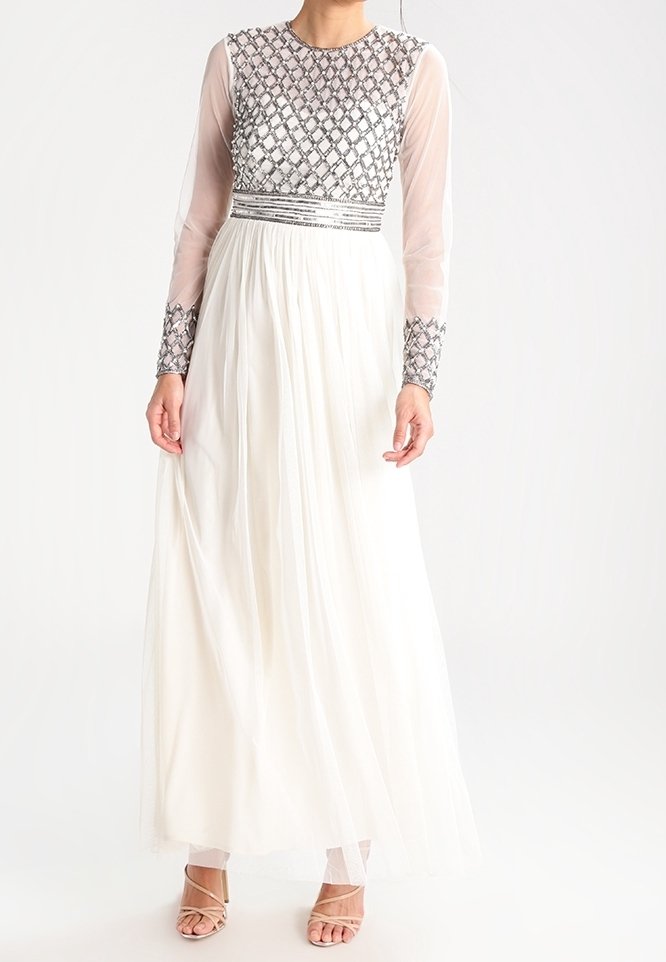 Robe longue blanche avec des manches longues transparentes, un corsage perlé et une jupe fluide. Présente un travail de perles complexe et un design texturé.