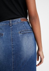 Džínová sukně v středním odstínu, s vysokým pasem, dvěma zadními kapsami a koženou značkovou nášivkou na pase. Hladká textura.