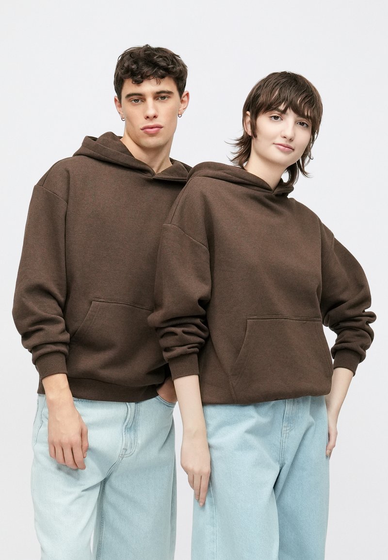 Braune Kapuzenpullover mit einer Fronttasche, kombiniert mit hellblauen Jeans. Der Stoff wirkt weich, mit lockerem Sitz und Bündchen.