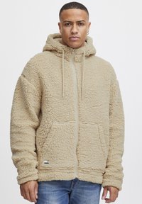 Beige fleece zip-up hette med en teksturert overflate, frontlommer og justerbare snorer på en romslig hette. Har et logo merke.
