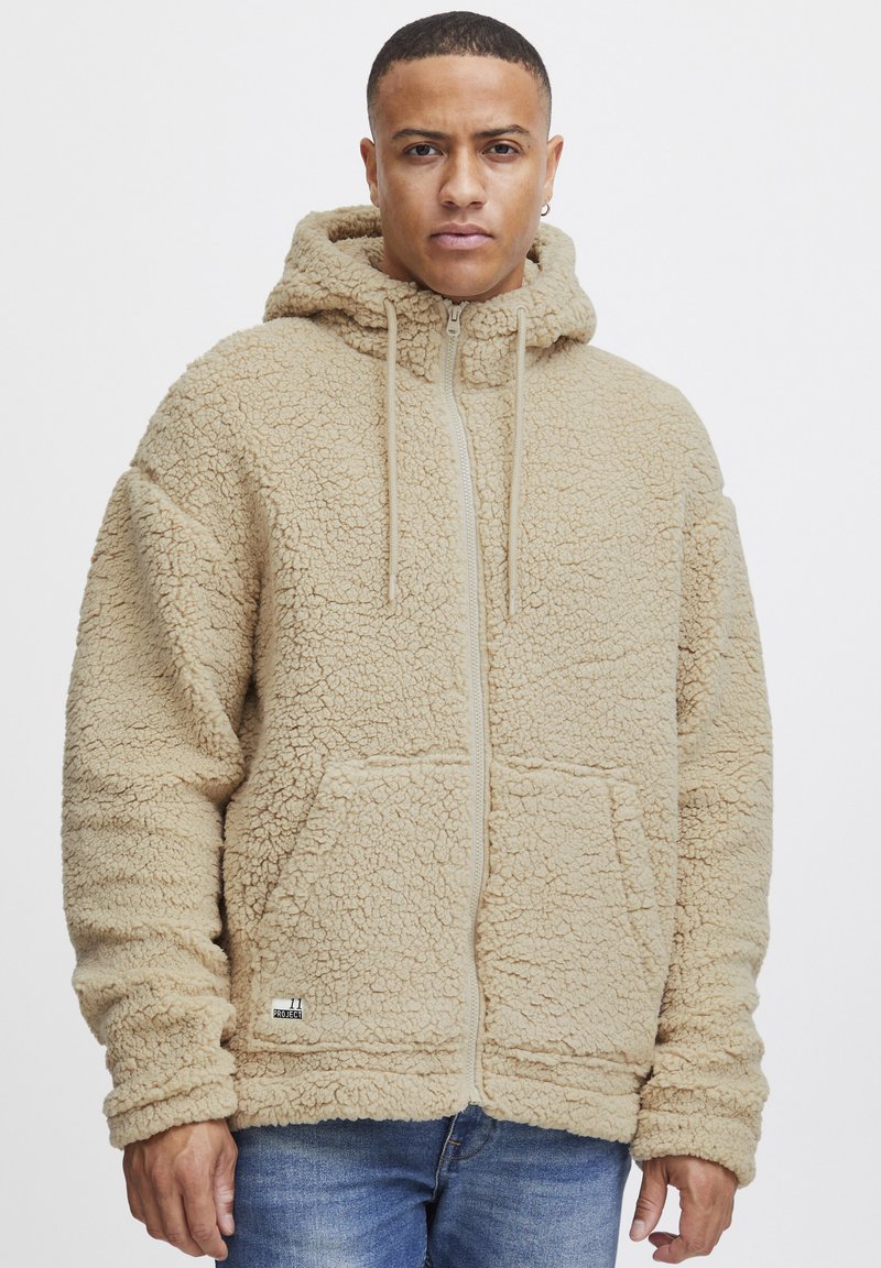 Beige fleece zip-up hette med en teksturert overflate, frontlommer og justerbare snorer på en romslig hette. Har et logo merke.