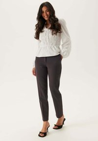 Witte blouse met lange mouwen en geplooid lijfje, gecombineerd met op maat gemaakte donkergrijze broek en zwarte sandalen met hak en strikdetail.