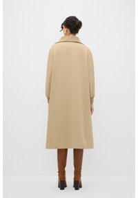 Trench beige con cuello tipo solapa, mangas largas y un corte holgado, combinado con botas de tacón marrones hasta la rodilla.