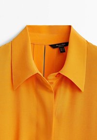 Chemise orange vif à col avec une patte de boutonnage cachée et une étiquette de marque visible à l'intérieur de l'encolure.