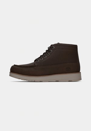 Bottines en daim marron avec une texture lisse, bout rond, œillets en métal et semelle en caoutchouc grise contrastante, arborant un logo à l'arrière.