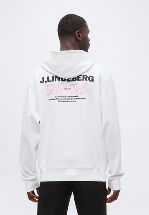 Mann mit dem Rücken zur Kamera, trägt einen weißen Hoodie mit schwarzem und pinkfarbenem Text, der das 30-jährige Jubiläum von J. Lindeberg von 1996 bis 2026 feiert.