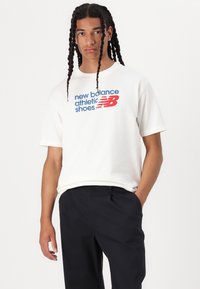 White cotton t-shirt featuring a blue and red graphic logo that reads "new balance athletic shoes." Paired with dark navy trousers.