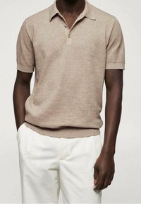 Homme portant un polo tricot léger marron clair à manches courtes et un pantalon blanc à plis, se tenant les mains dans les poches.