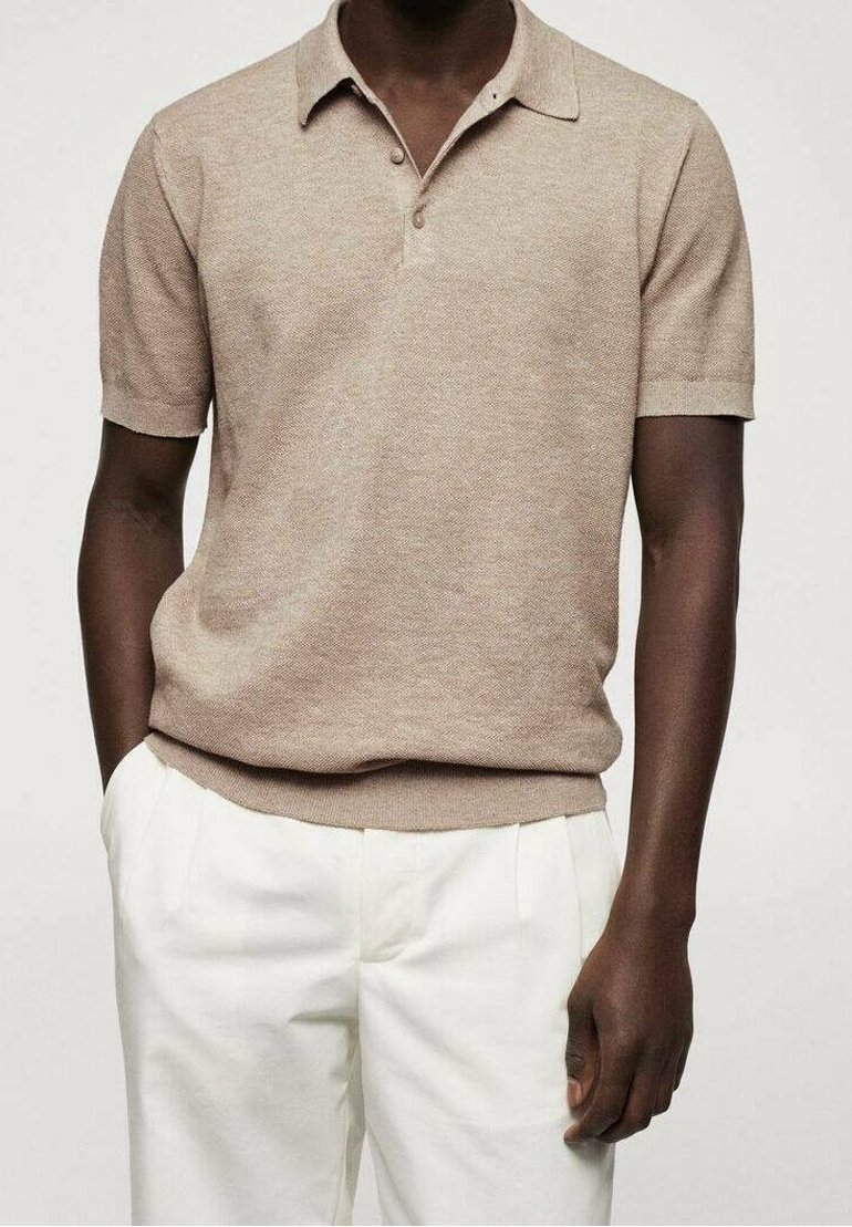 Homme portant un polo tricot léger marron clair à manches courtes et un pantalon blanc à plis, se tenant les mains dans les poches.