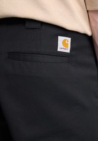 Czarne spodnie z mocnego materiału, z boczną kieszenią i kwadratową metką Carhartt w kolorze żółtym i białym.