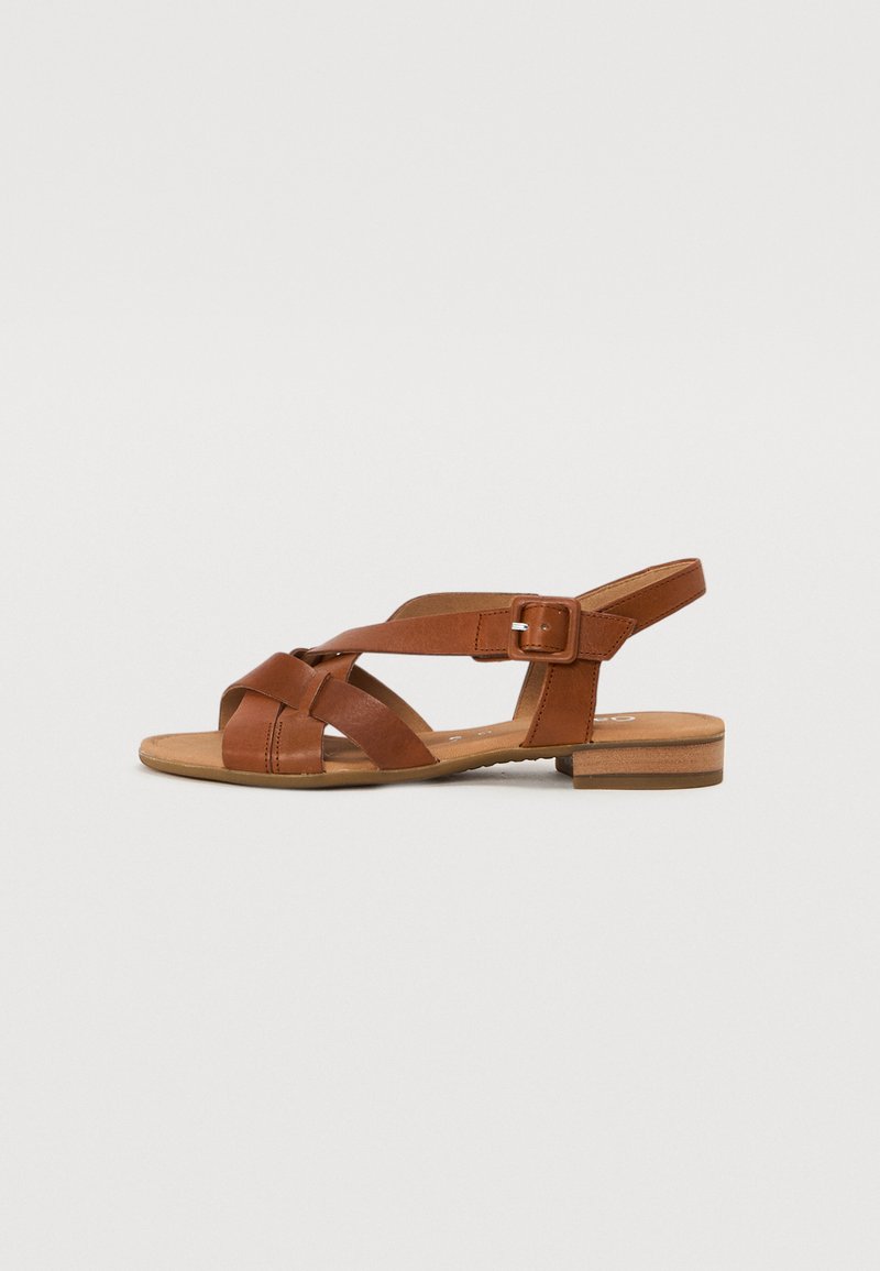 Brune lædersandal med flad sål, der har krydsende stropper og spænde lukning ved anklen. Glat tekstur, afslappet design.