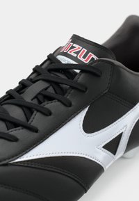 Crne kožne nogometne kopačke s bijelim detaljima, glatkom teksturom i crnim vezicama. Značajna karakteristika uključuje logo na jeziku.