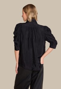 Svart bluse med en strukturert bakside, puffermer og en glatt, shiny tekstur. Har høy krage og knapper på ermene.