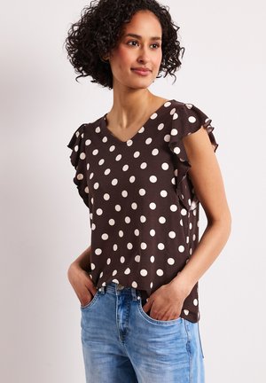 Kobieta o kręconych włosach, ubrana w brązową bluzkę w białe kropki z falbankowymi rękawami oraz niebieskie dżinsy, ręce w kieszeniach, patrzy w prawo.