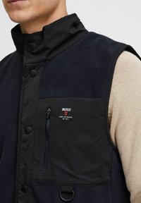 Chaleco negro y azul marino con cuello alto, botones a presión, textura de forro polar, bolsillo con cremallera y etiqueta con logotipo en detalles rojos, blancos y negros.