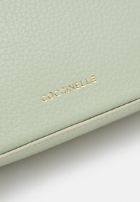 Svetlo zelena teksturirana usnjena torbica z embosiranim zlatim logotipom "Coccinelle", ki vključuje gladek rob in podrobnosti šivanja.