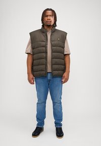 Olivovo zelená vypchatá vesta s vysokým golierom, nosená cez béžové tričko. V páre s modrými džínami a čiernymi teniskami. Diskrétne logo na veste.