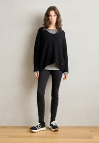 Schwarzer übergroßer Pullover mit V-Ausschnitt, getragen über einem gestreiften Hemd, kombiniert mit schwarzen Skinny Jeans und schwarzen Sportschuhen mit weißen Akzenten.