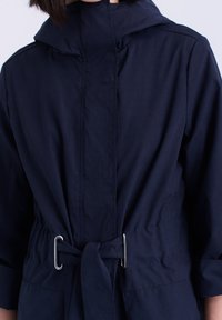 Veste légère bleu marine avec capuche, taille ceinturée avec quincaillerie argentée. Tissu texturé avec détails de coutures visibles.