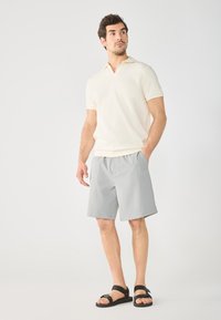 Polo shirt beige clair à manches courtes avec une texture côtelée et un col V, associé à un short gris et des sandales noires à straps réglables.