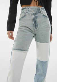 Pantalones vaqueros de mezclilla con un diseño de talle alto, en un tono azul claro desgastado y paneles de tela blanca en contraste. Los dobladillos deshilachados añaden detalle.