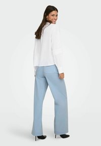 Femme souriante regardant par-dessus son épaule, portant un chemisier blanc à manches longues avec des poignets en dentelle, un pantalon large bleu clair et des chaussures noires à talons hauts.