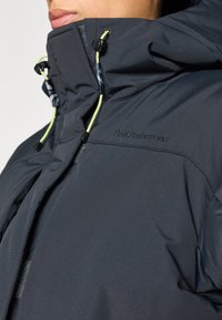 Tumesinine polsterdatud jakk kõrge kraega, kontrastse neoonroheliste paeladega ja musta logoga rinnal. Sileda tekstuuriga kangas tikitud mustriga.