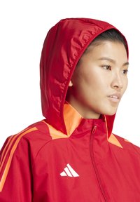 Veste de sport rouge avec capuche, fermeture éclair et accents orange. Fabriquée dans un matériau lisse et léger avec trois bandes blanches de marque.
