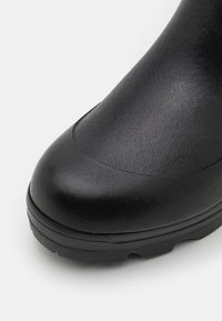 Botte en caoutchouc noir avec une surface texturée, un bout arrondi et une semelle épaisse à crampons pour une meilleure adhérence. Aucun matériel ou accent visible.