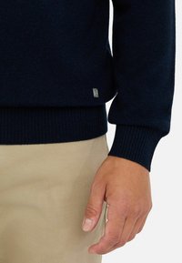Granatowy sweter z dzianiny z prążkowanymi mankietami i dołem, z małym metalicznym logo po boku, noszony na beżowych spodniach.