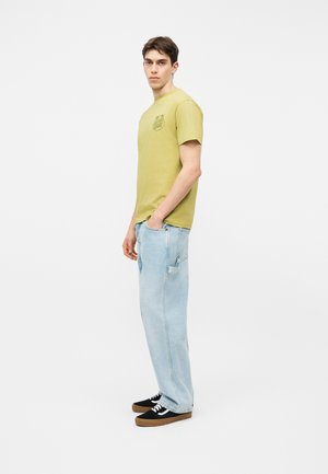 Fiatal férfi oldalra fordulva, kezét a zsebében tartva, világoszöld pólót, laza világoskék farmert és fekete sportcipőt barna talppal visel.