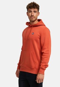 Ung mann med kort krøllete hår iført en brent oransje hettegenser med frontlomme og svarte bukser, stående foran en ensfarget hvit bakgrunn.