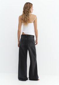Femme aux longs cheveux ondulés, vue de dos, portant un haut blanc à fines bretelles et un jean noir large et ample sur fond blanc.