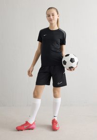 Must musta Nike treeningpluus ja lühikesed püksid valgete detailidega, roosade jalgpalliketsade ja põlveni valgete sokkidega, hoides musta-valget jalgpalli.