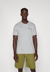 Grå champion T-shirt med korte ærmer og rund halsudskæring, der har et lille sort logo på brystet. Sæt med olivengrønne shorts.