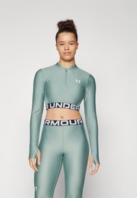 Žalias sportinis trumpas viršus su ilgomis rankovėmis, užtrauktuku prie kaklo, derinamas su atitinkančiomis legginsomis, turinčiomis juodą juostą su užrašu "UNDER ARMOUR".