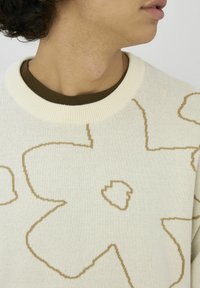 Gros plan sur le cou et la partie inférieure du visage d'une personne portant un pull crème avec un grand motif floral beige sur une chemise marron.