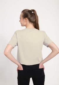 Helles beige T-Shirt mit kurzen Ärmeln und Rundhalsausschnitt, aus weichem Stoff. Modell von hinten gezeigt, Hände in den Taschen, mit dunklen Jeans.