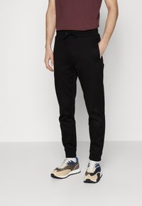 Black sweatpants with a slim fit, drawstring waist, and side pockets; paired with beige, blue, and navy athletic shoes.