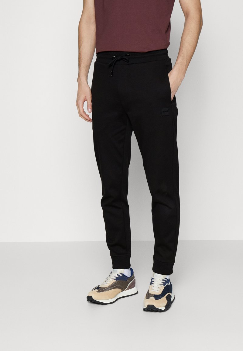 Black sweatpants with a slim fit, drawstring waist, and side pockets; paired with beige, blue, and navy athletic shoes.