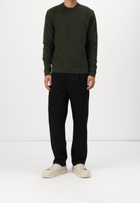 Olivgrön stickad tröja med ribbad nederkant, parat med löst sittande svarta byxor. Skorna är krämfärgade sneakers med texturerade sulor.