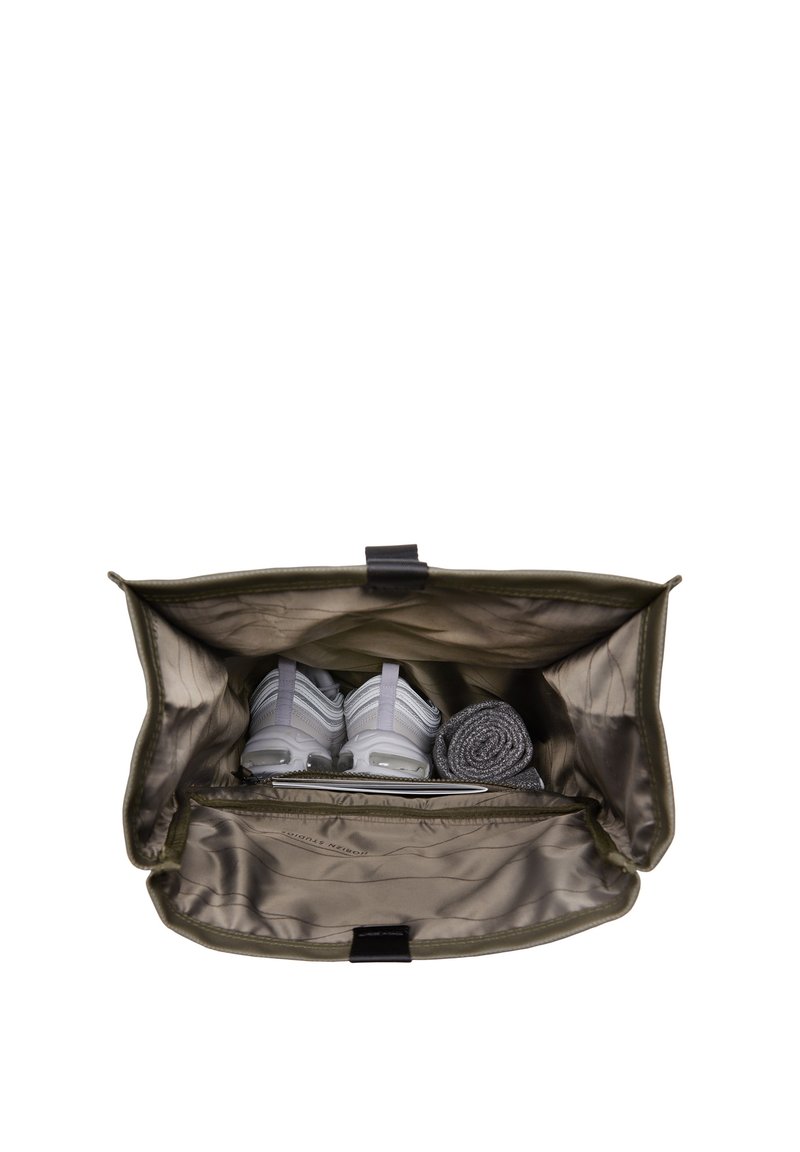 Sac en tissu vert avec un intérieur argenté, contenant des chaussures de sport grises, une serviette roulée et un objet plat dans un compartiment zippé.