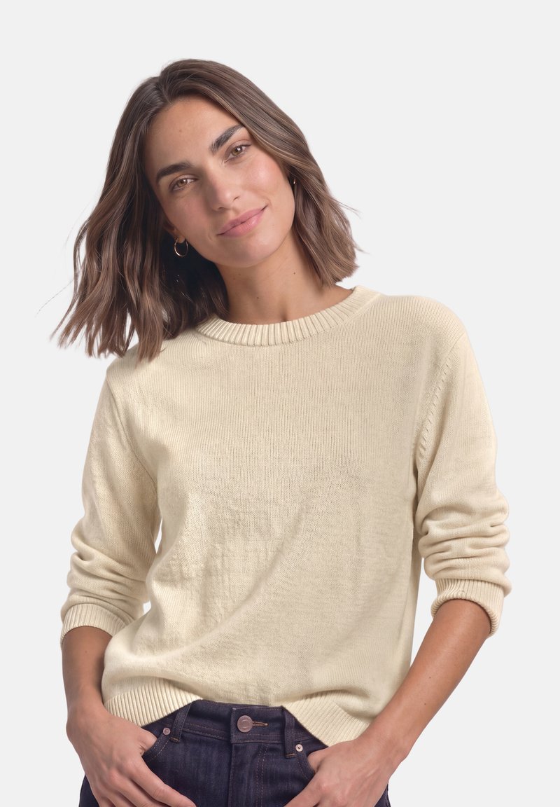 Femme aux cheveux bruns mi-longs portant un pull crème et un jean foncé, souriante les mains dans les poches sur un fond uni.